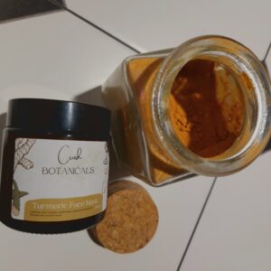 Jar of natural turmeric clay face mask laid down on white tiles, next to an open jar of turmeric powder.