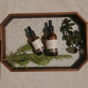 Bottles of scalp oil for hair growth on glass tray surrounded by rosemary and peppermint leaves on a beige backround